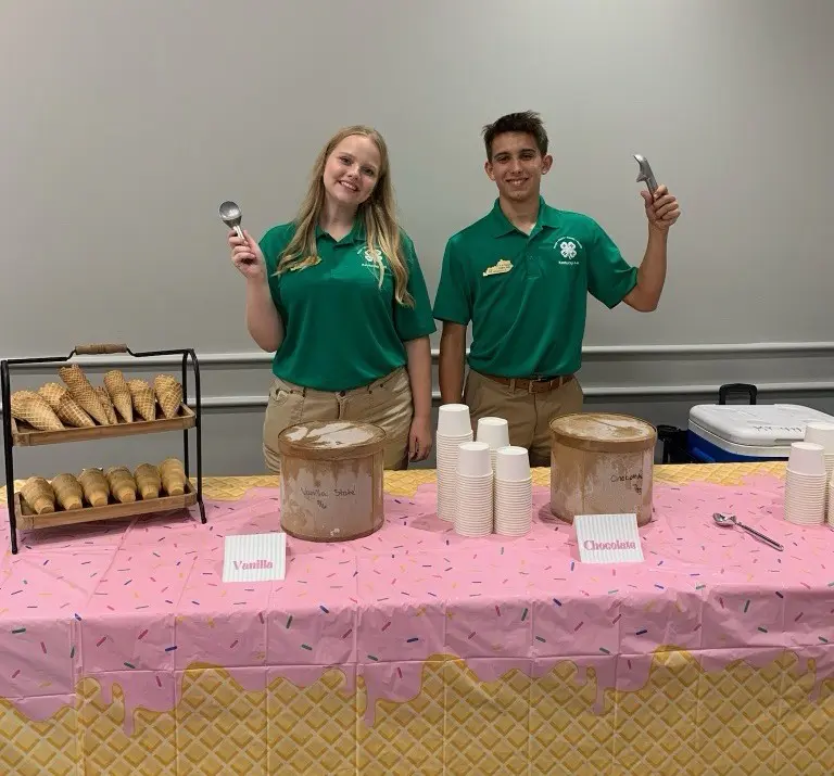 Two 4-H State officers stand behind an ice cream social table, smiling and holding ice cream scoops. The table is decorated with an ice cream-themed tablecloth and features waffle cones, cups, and tubs of vanilla and chocolate ice cream ready to serve.