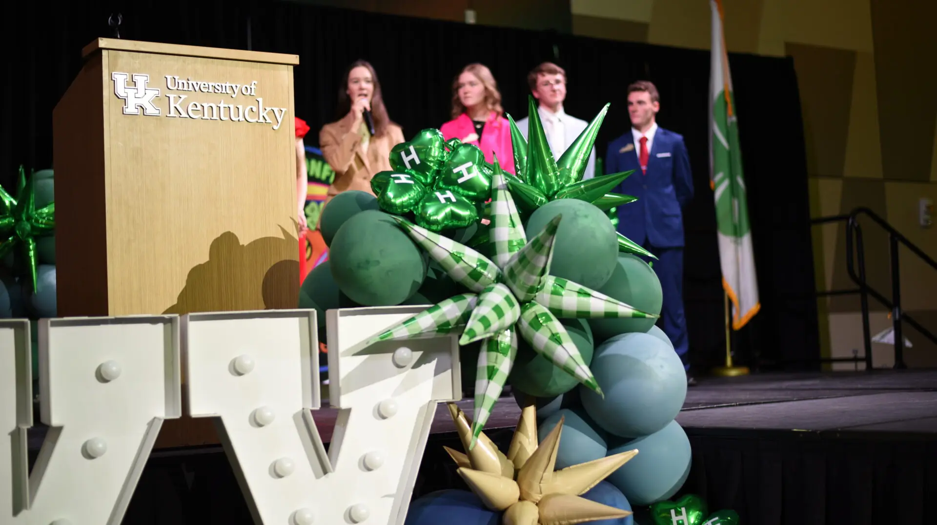 4-H balloons with 4-H youth in the background on a stage.